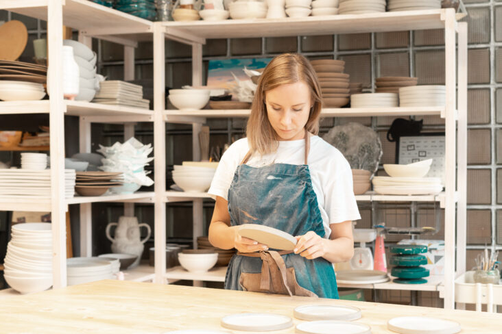 jovem mulher junto a uma bancada de cerâmica , tendo por detras uma estante com várias peças que produziu no seu trabalho extra