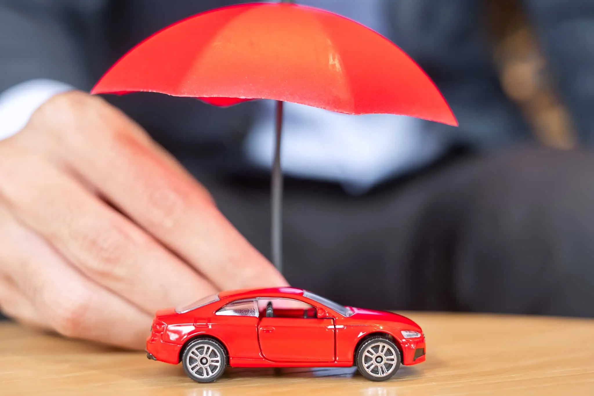 Sabe verificar o seguro pela matrícula do seu automóvel? mão de homem a segurar um chapeu de chuva em miniatura, que está a cobrir um automóvel tambbém miniatura, ambos vermelhos, representando a possibilidade de verificar seguro pela matrícula