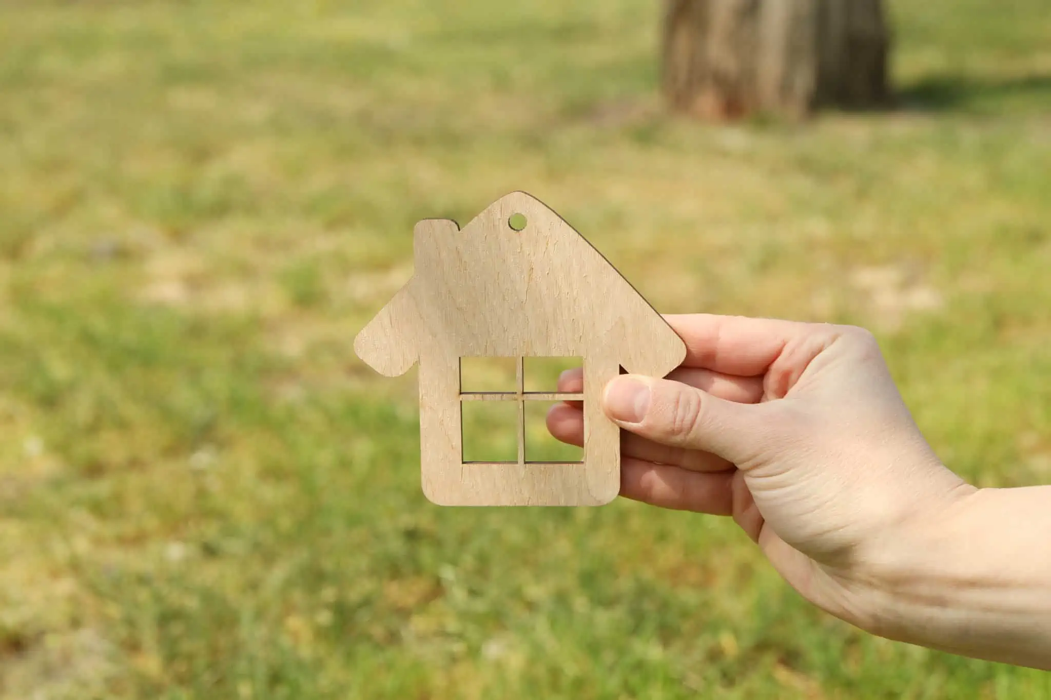 Quero arrendar a minha casa e comprar outra. Como funciona? Mão a segurar casa de madeira com relva por trás simbolizando arrendar e comprar casa
