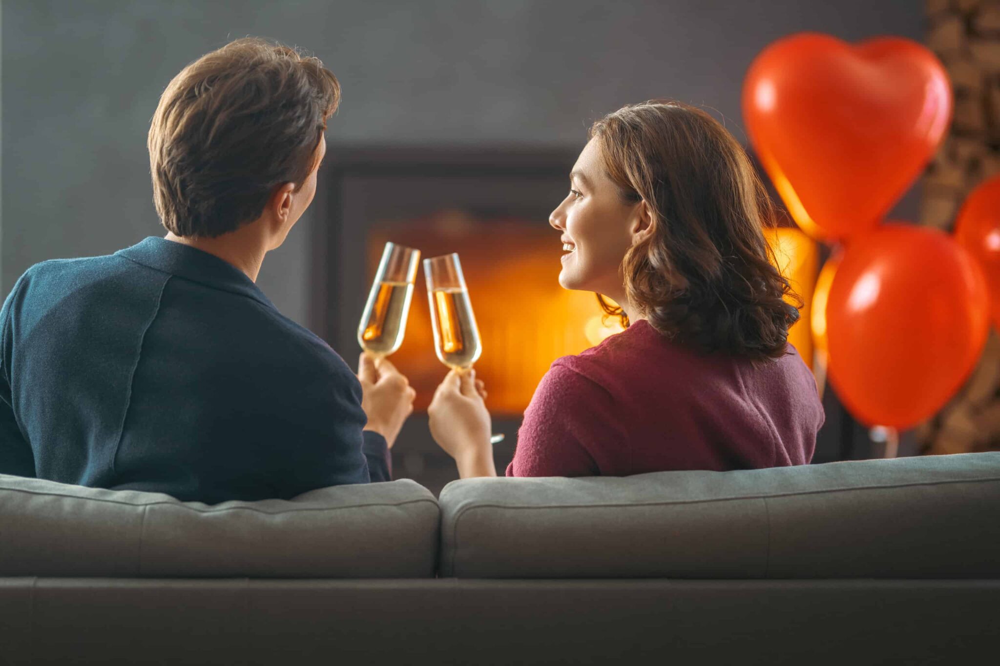 casal sentados no sofa da sala frente à lareira, a brindar e com balões vermelhos a celebrar o dia dos Namorados