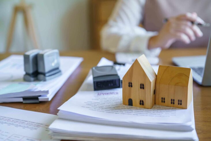 em fundo, desfocado, uma mulher segura caneta para assinar documentos, empilhados na mesa com miniaturas de imóveis em cima, sendo que a reserva de propriedade aplica-se tanto a crédito para casa ou carro