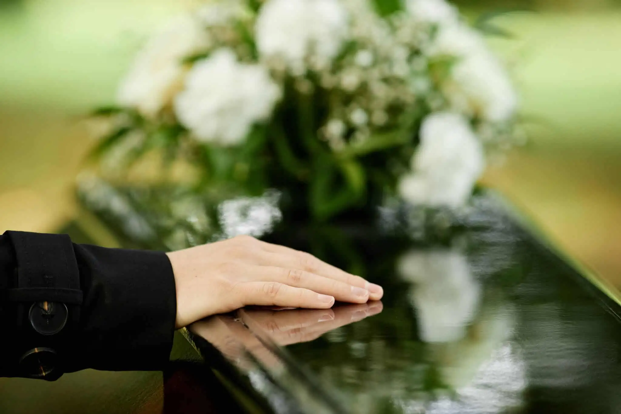 Subsídio de funeral: Quem tem direito e como requerer? mão de jovem mulher repousa sob um caixão de madeira escura, com um ramo de flores desfocado em fundo, ilustrando o subsídio de funeral