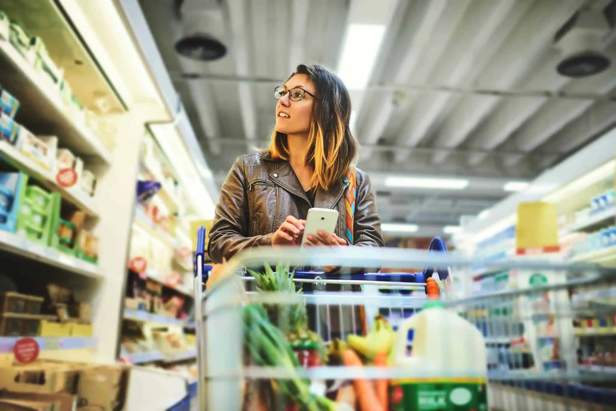 jovem mulher no corredor do supermercado empurra o carrrinho de comprs qu decide com o telemóvel na mão enquanto consulta 5 sites de vales de desconto para supermercado e outros em Portugal