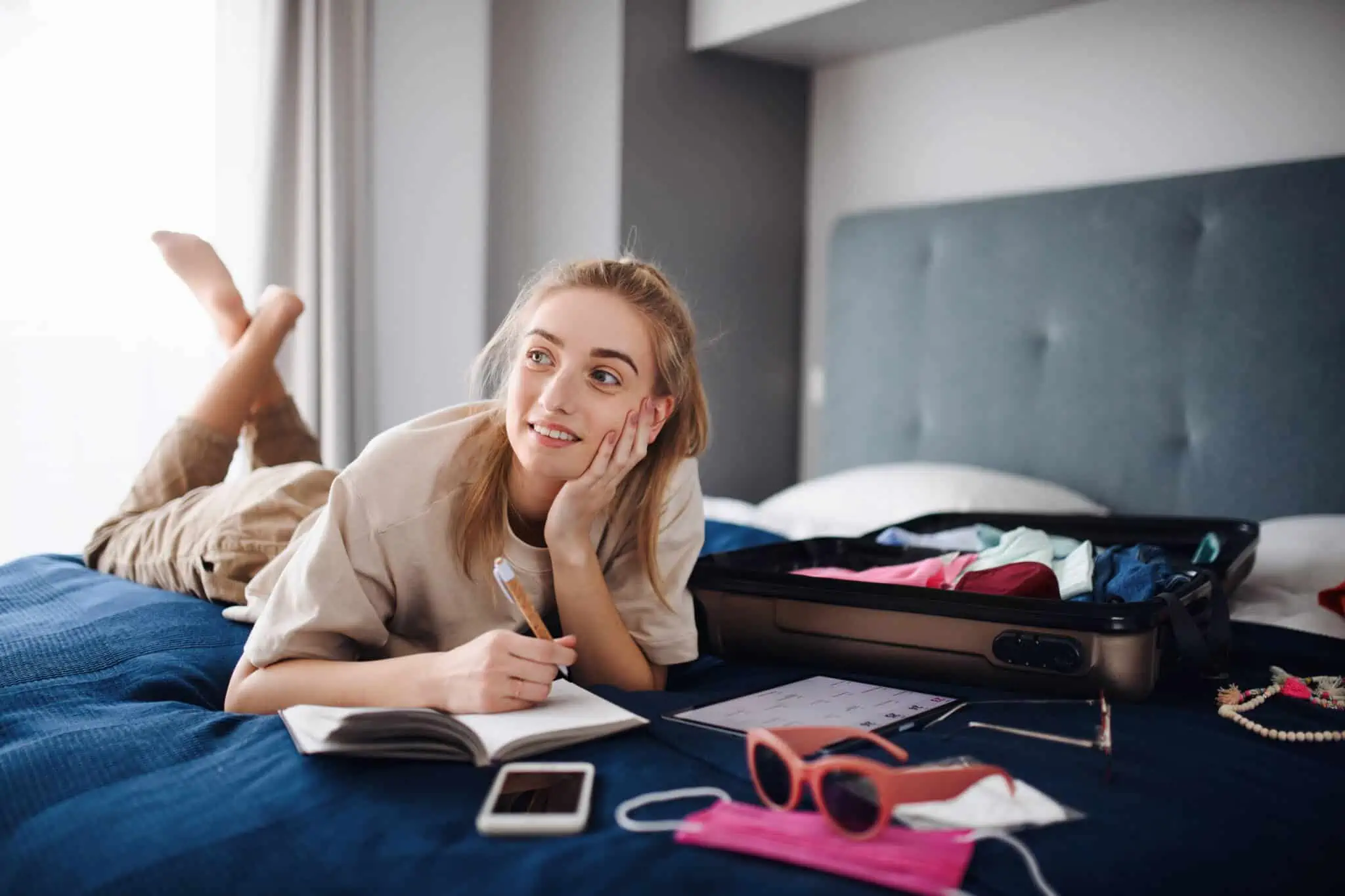 Mapa de férias: O que diz o Código do Trabalho jovem mulher deitada na sua cama, onde tem a mala e vários objetos de viagem, enquanto planeia como poupar nas férias