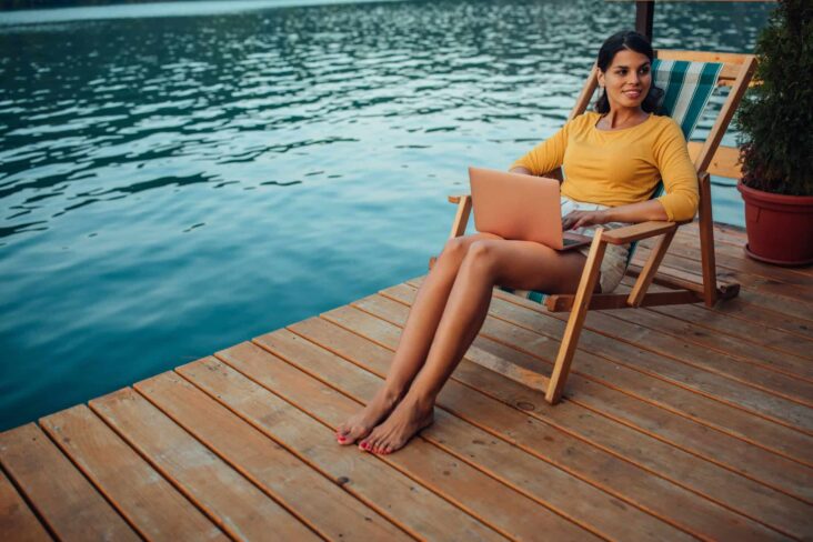 jovem mulher sentada numa cadeira de madeira, sob um decke de madeira, ao redor de uma piscina, com o computador portátil no colo a preparar as férias com antecedência