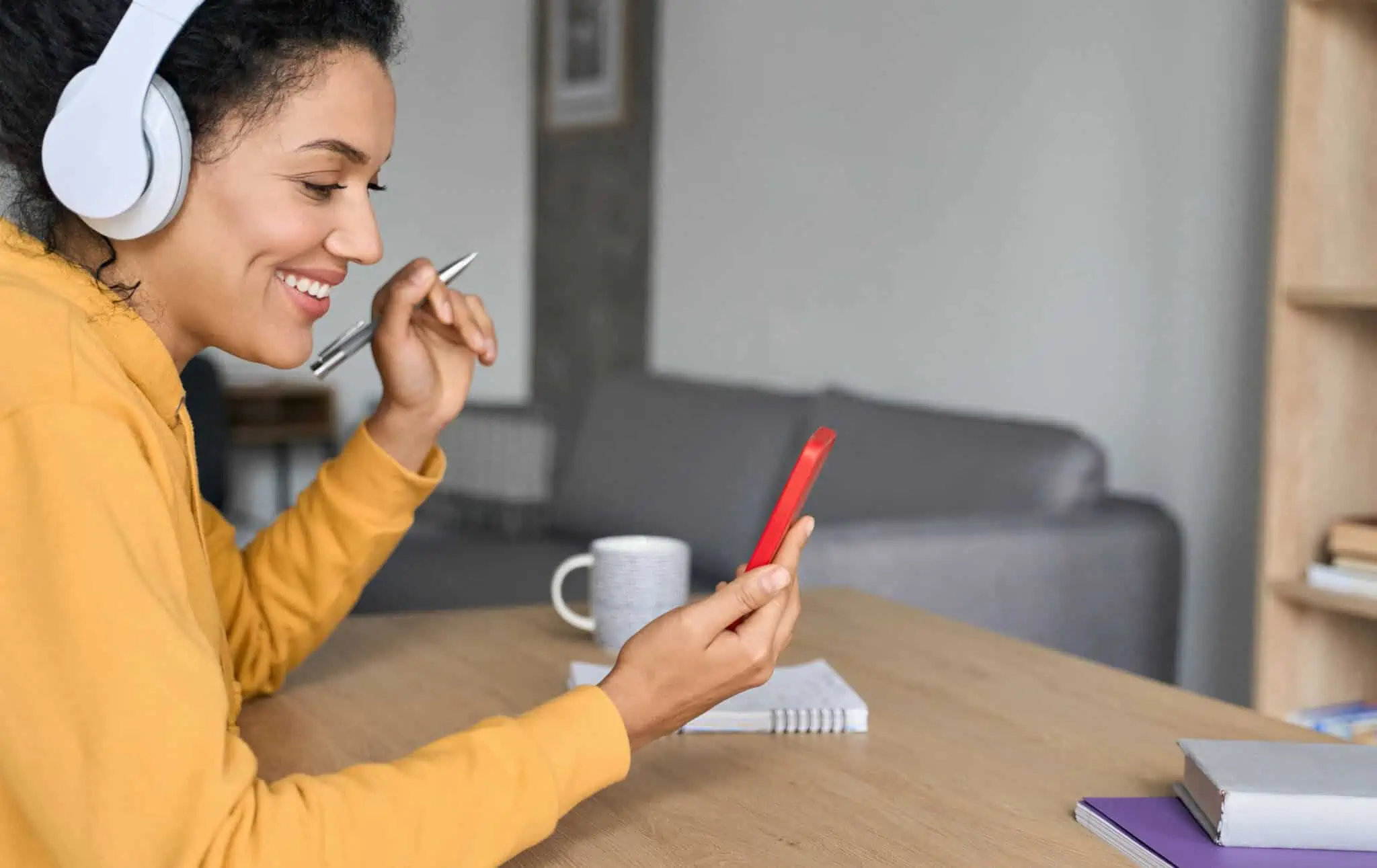 15 sites para aprender inglês online e outras sugestões jovem mulher em casa, sentada à mesa da sala, ao telemóvel usa aplicação para aprender inglês, tirando notas para um bloco de papel
