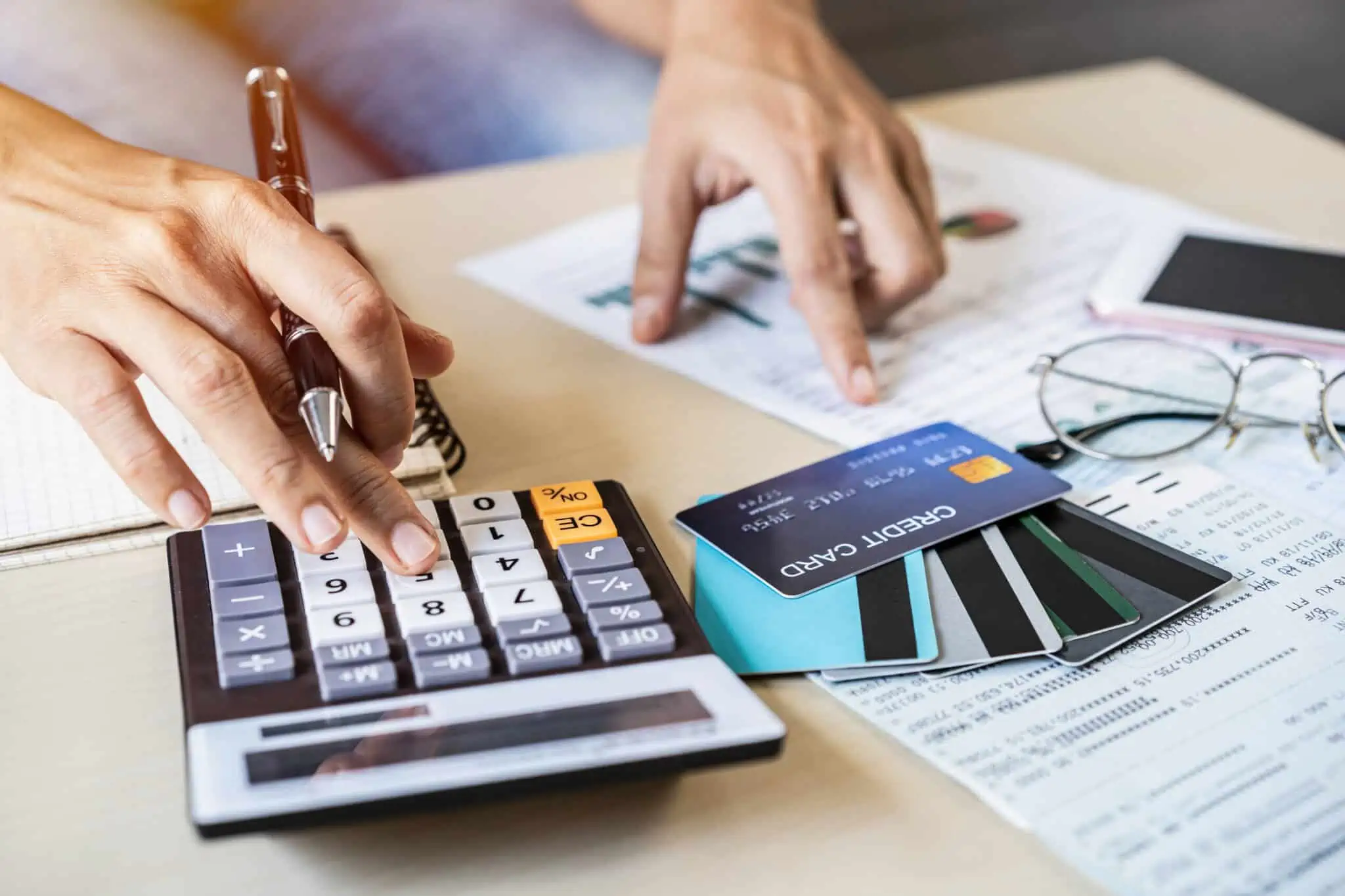 Preparado para escolher a “melhor” conta bancária para si? mão de jovem mulher a fazer contas na calculadora e analisar documentos espalhados na mesa onde estão também um conjunto de cartões bancários, produtos de contas bancárias