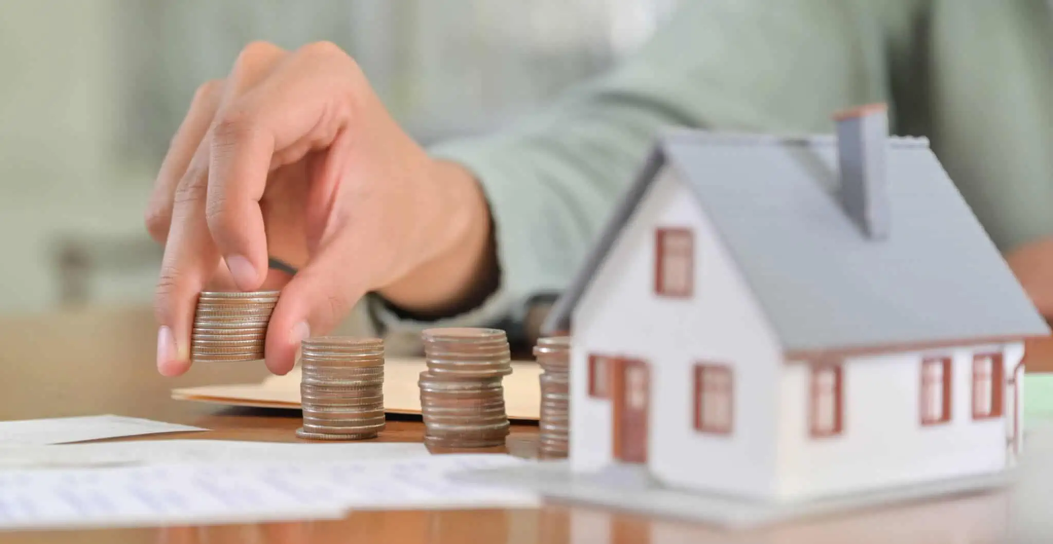 Quanto custa fazer um crédito habitação? uma casa, com uma pessoa a colocar moedas ao lado e papeis na mesa a ilustrar quanto custa fazer um crédito habitação