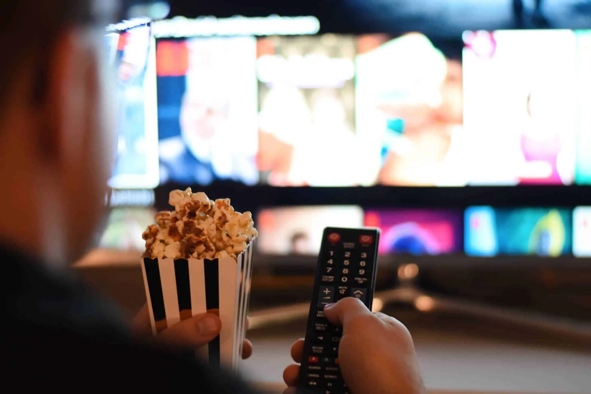 Educação financeira: Estes filmes podem aumentar os seus conhecimentos frente a uma televisão, com ecran desfocado, a mão de um jovem rapaz segura comando e pipocas, pronto para ver filmes que ajudam na educação financeira