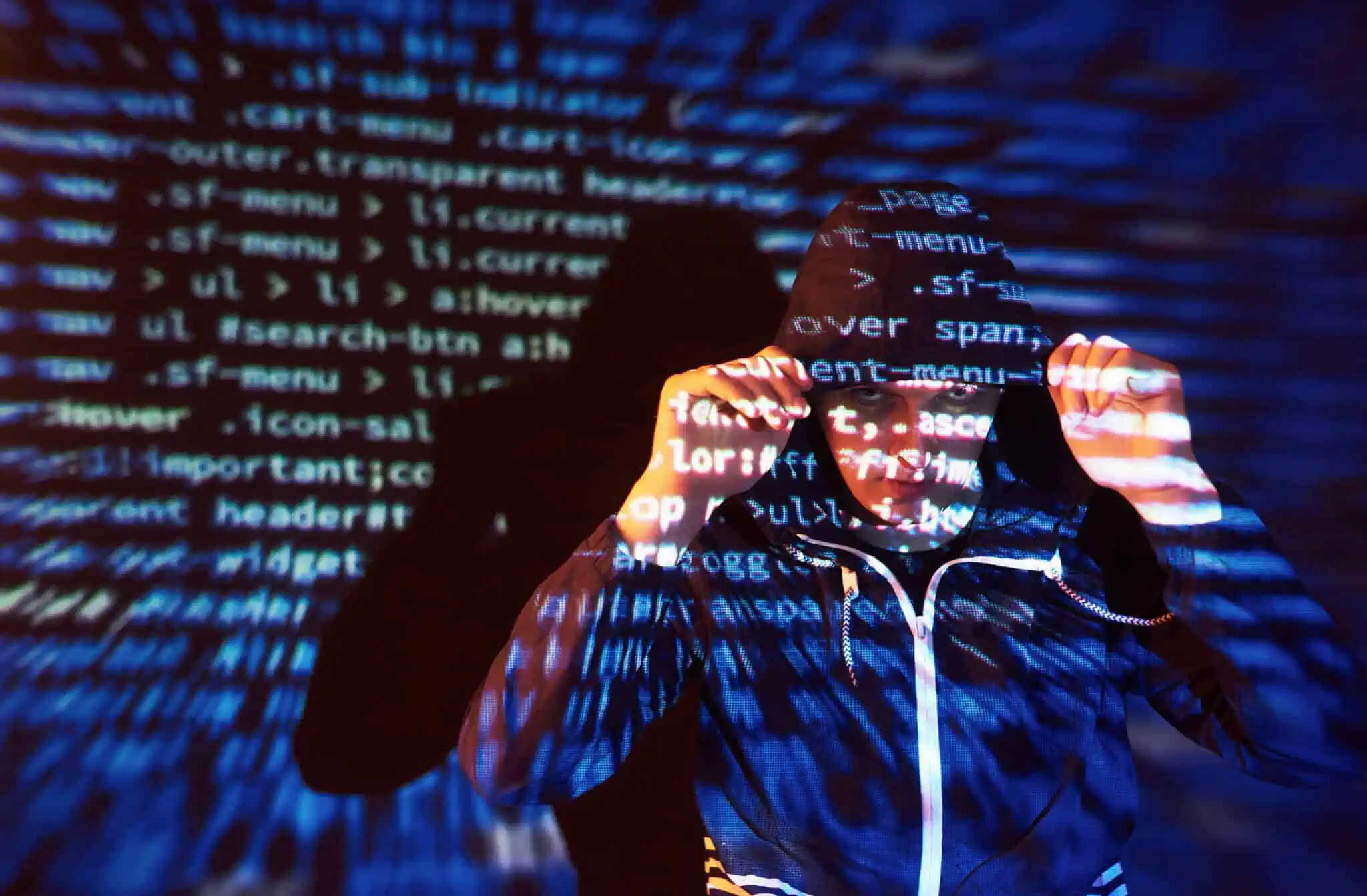 Roubo de identidade: O que é e o que deve fazer se for vítima jovem rapaz tapa a cabeça com capuz esta frente a projeções de códigos de programação que permitem o roubo de identidade