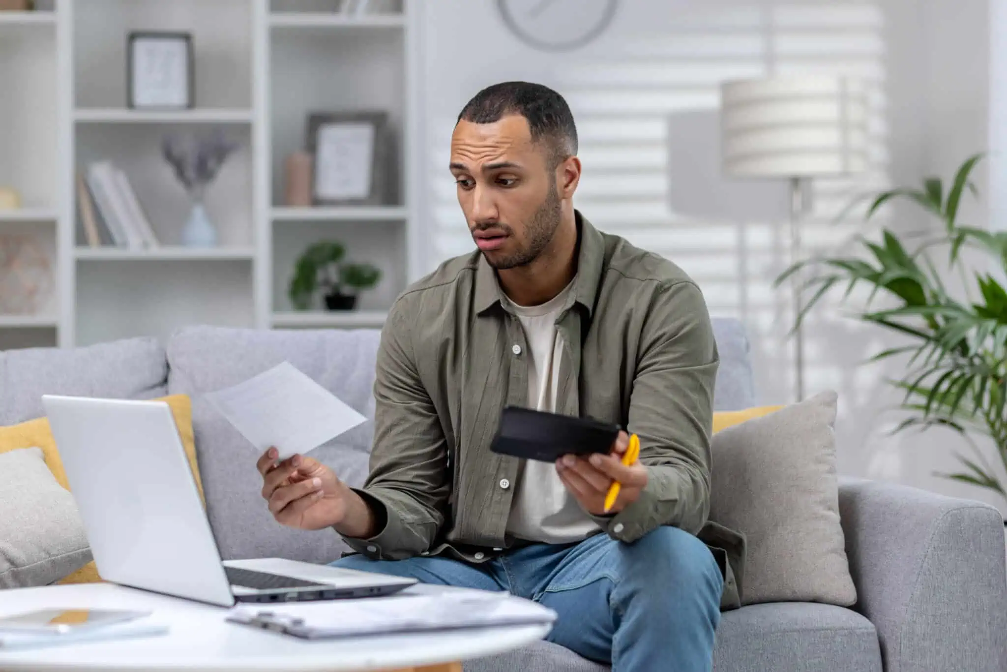 jovem homem a trabalhar na sua sala, ao computador, segura calculadora e documentos enquanto tenta perceber a gestão de responsabilidades do trabalho extra ou part-time
