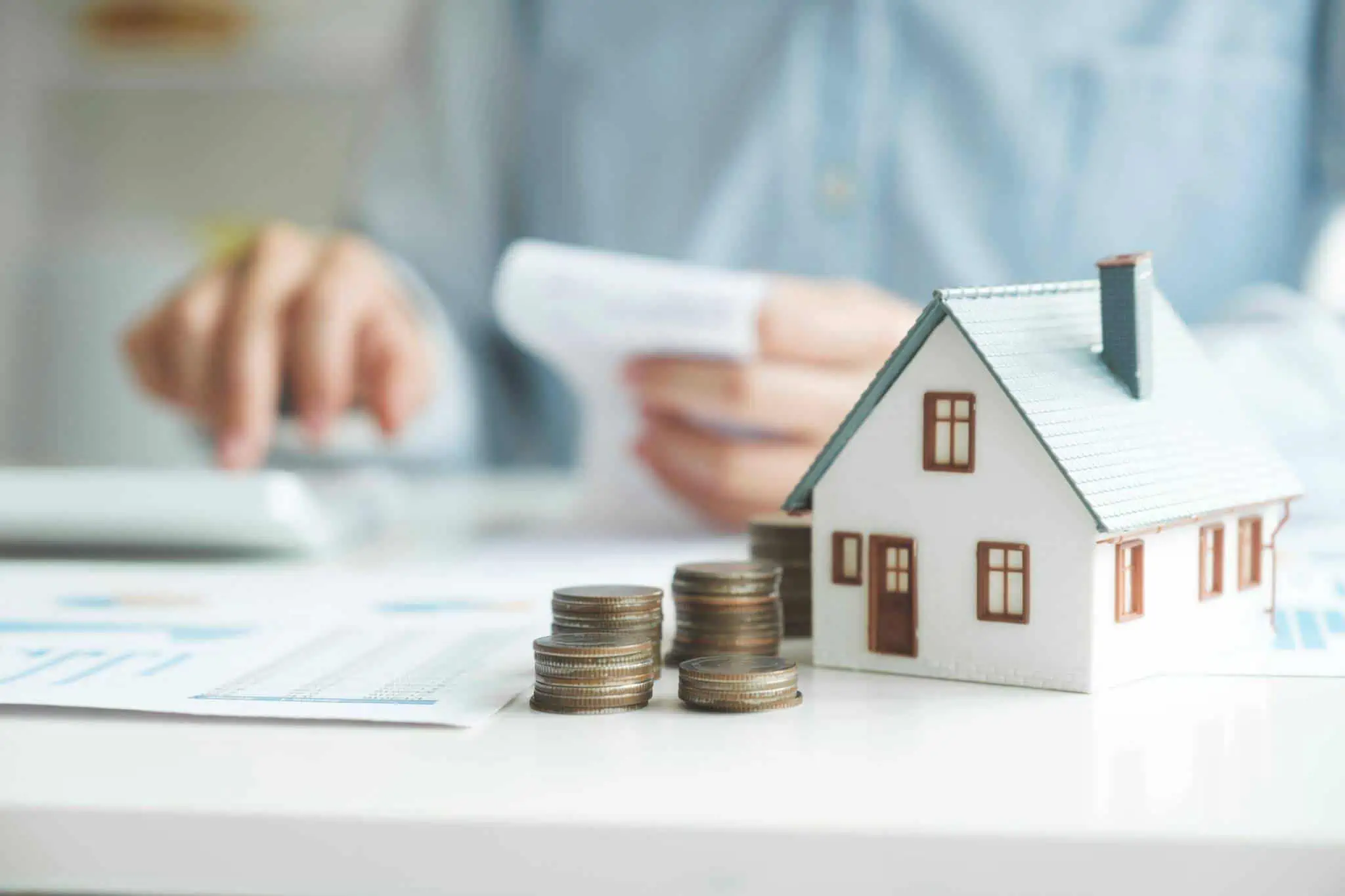 Qual o limite de financiamento no crédito habitação? casa pequena em cima da mesa com moedas ao lado. Ao fundo, um homem faz contas numa calculadora a ilustrar quando pagar o IMI em 2023