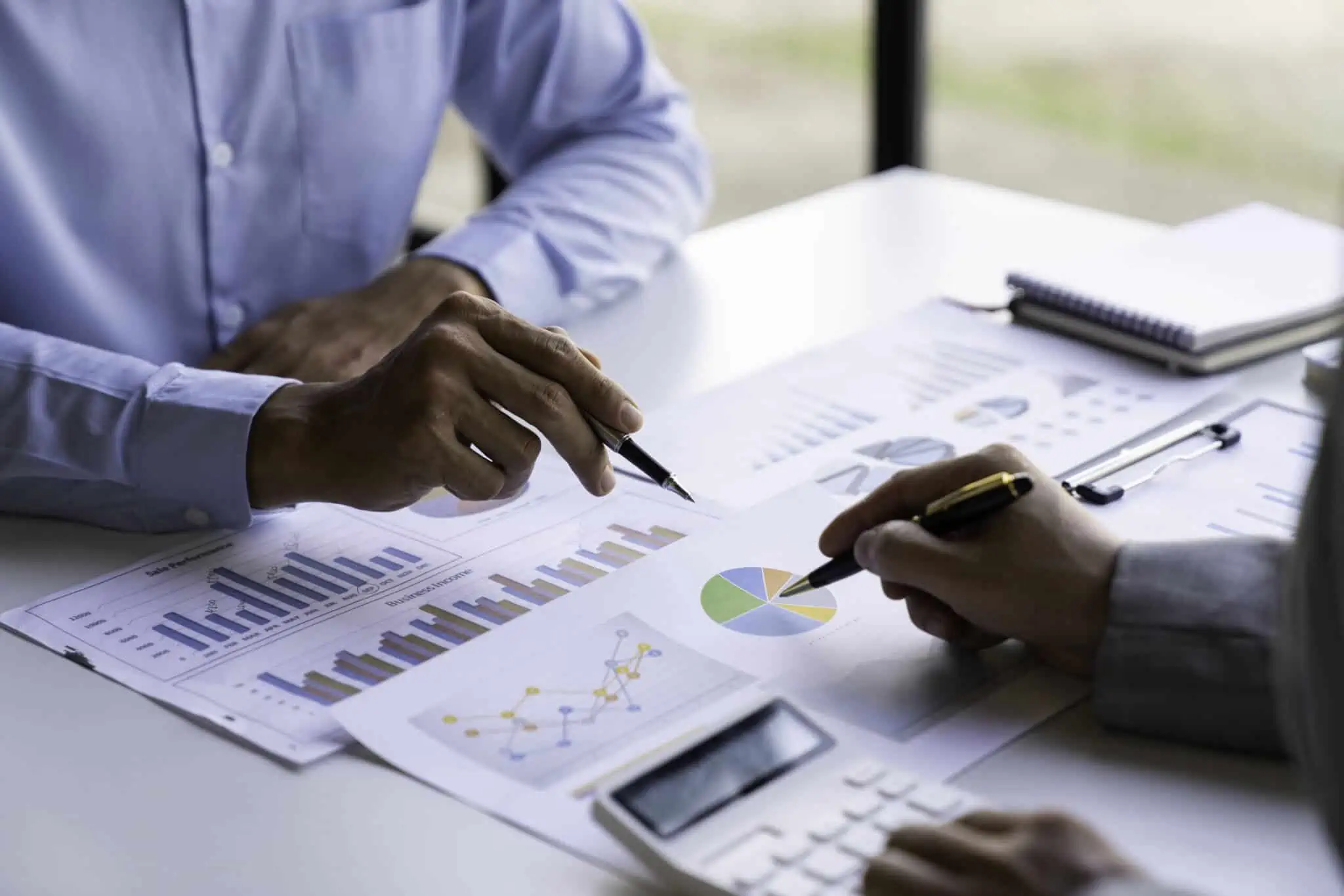 O que é a Informação Empresarial Simplificada? mesa coberta de documentos com gráficos e números que estão a ser calculados por dois homens com utilização da calculadora numa avaliação de empresa