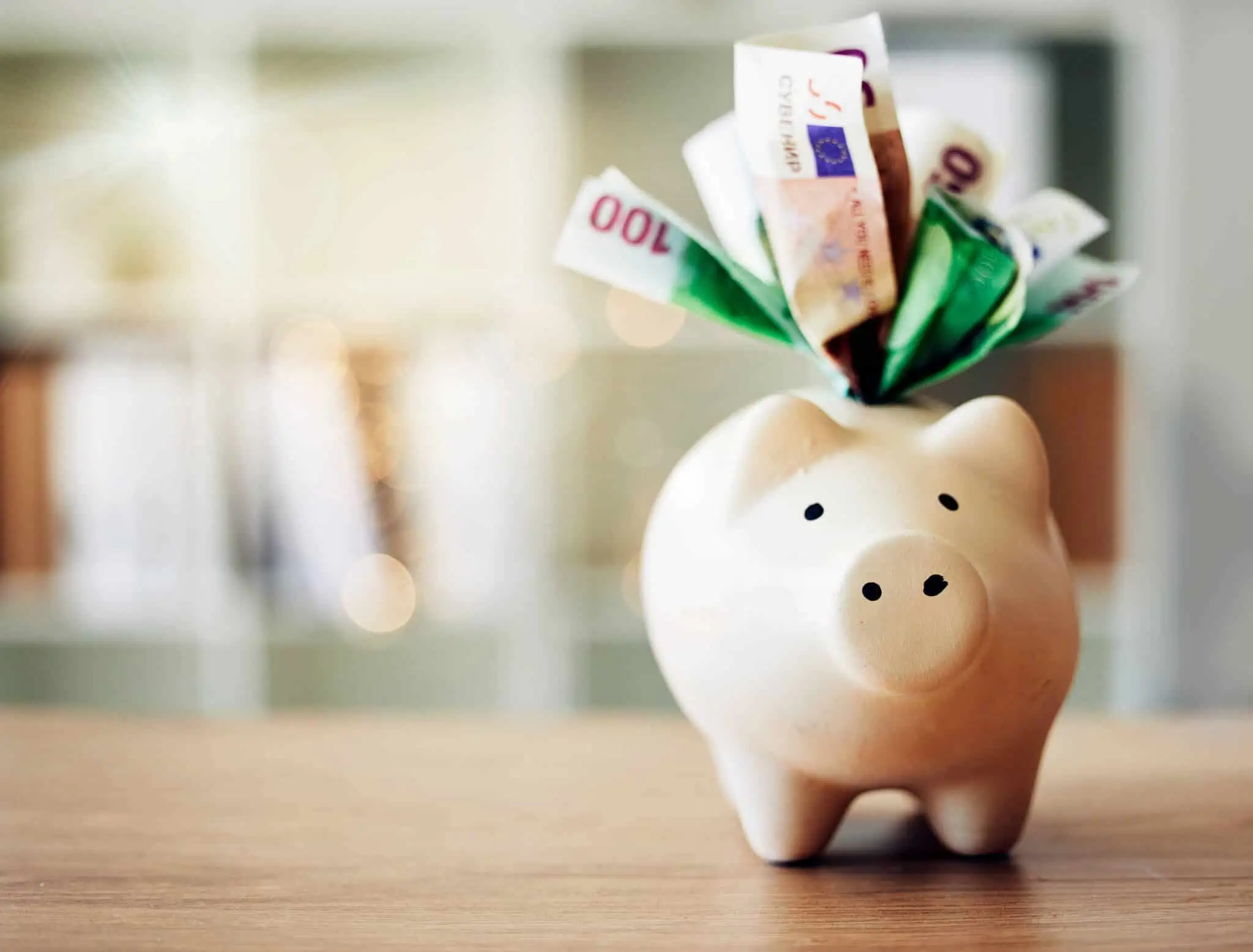 em fundo desfocado um porquinho mealheiro beje está em cima de uma mesa de madeira, clara, a receber algumas notas de diferentes valores de euros, as poupanças que assentam em descontos voluntários para a reforma