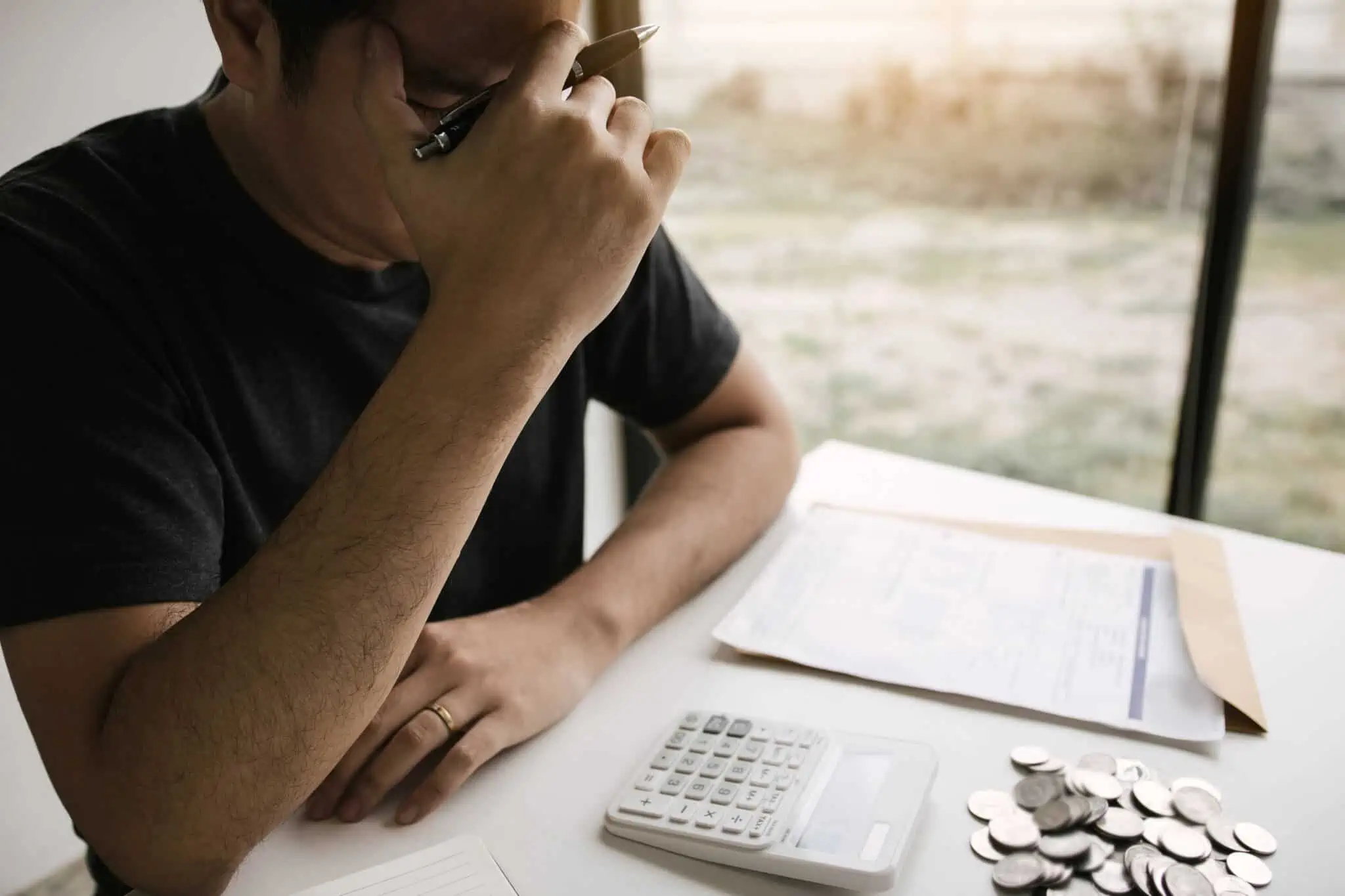 Simulador: Qual o nível de stress financeiro dos seus colaboradores? jovem rapaz a fazer contas com a calculadora, caneta e muitos papeis espalhados numa mesa, junto a uma janela, está com a mão na cabeça mostrando stress financeiro, o qual pode ser medido num simulador