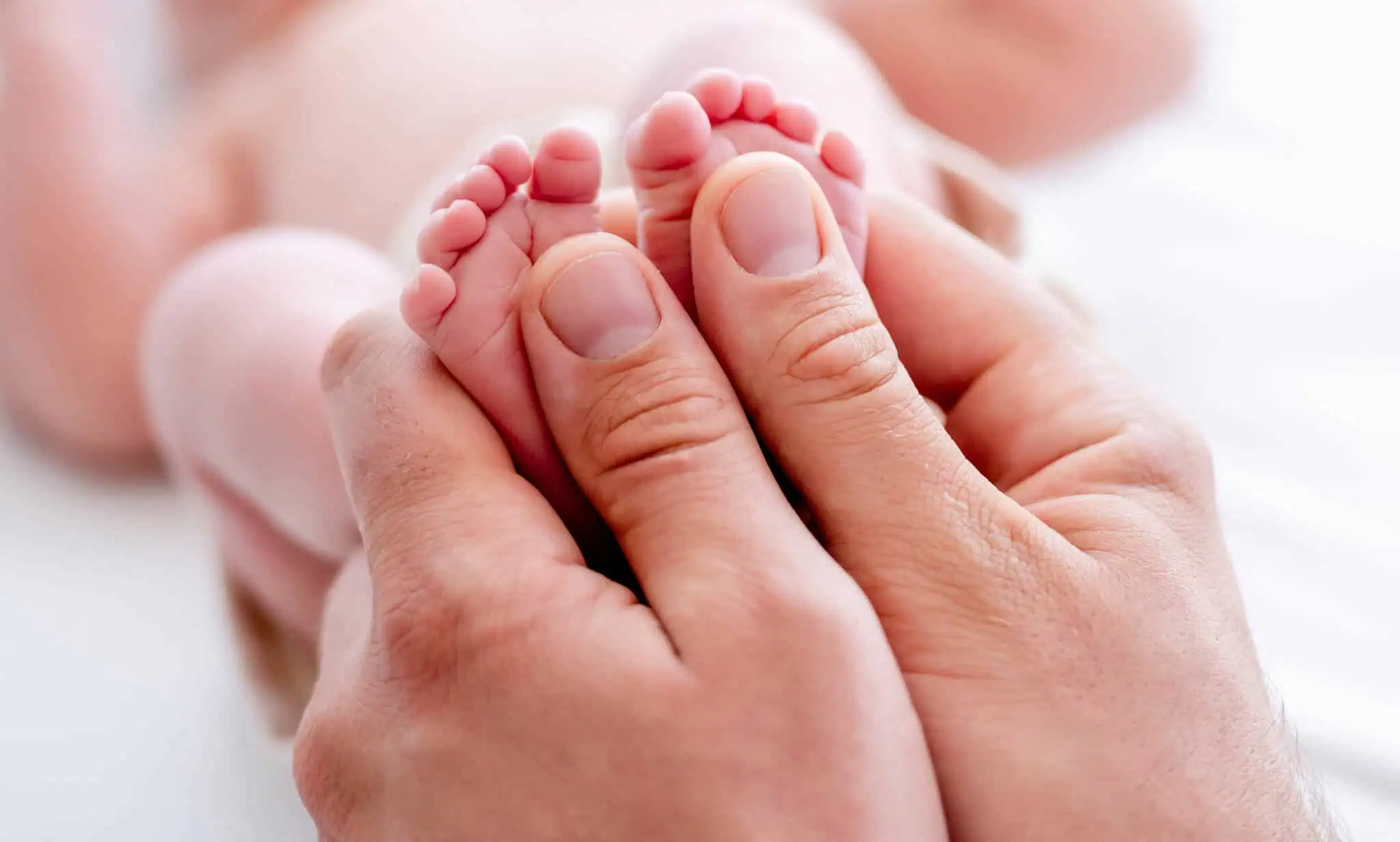 Licença de maternidade e paternidade: Como funcionam? mãos agarram pés de recém nascido deitado de barriga para cima, em fundo desfocado