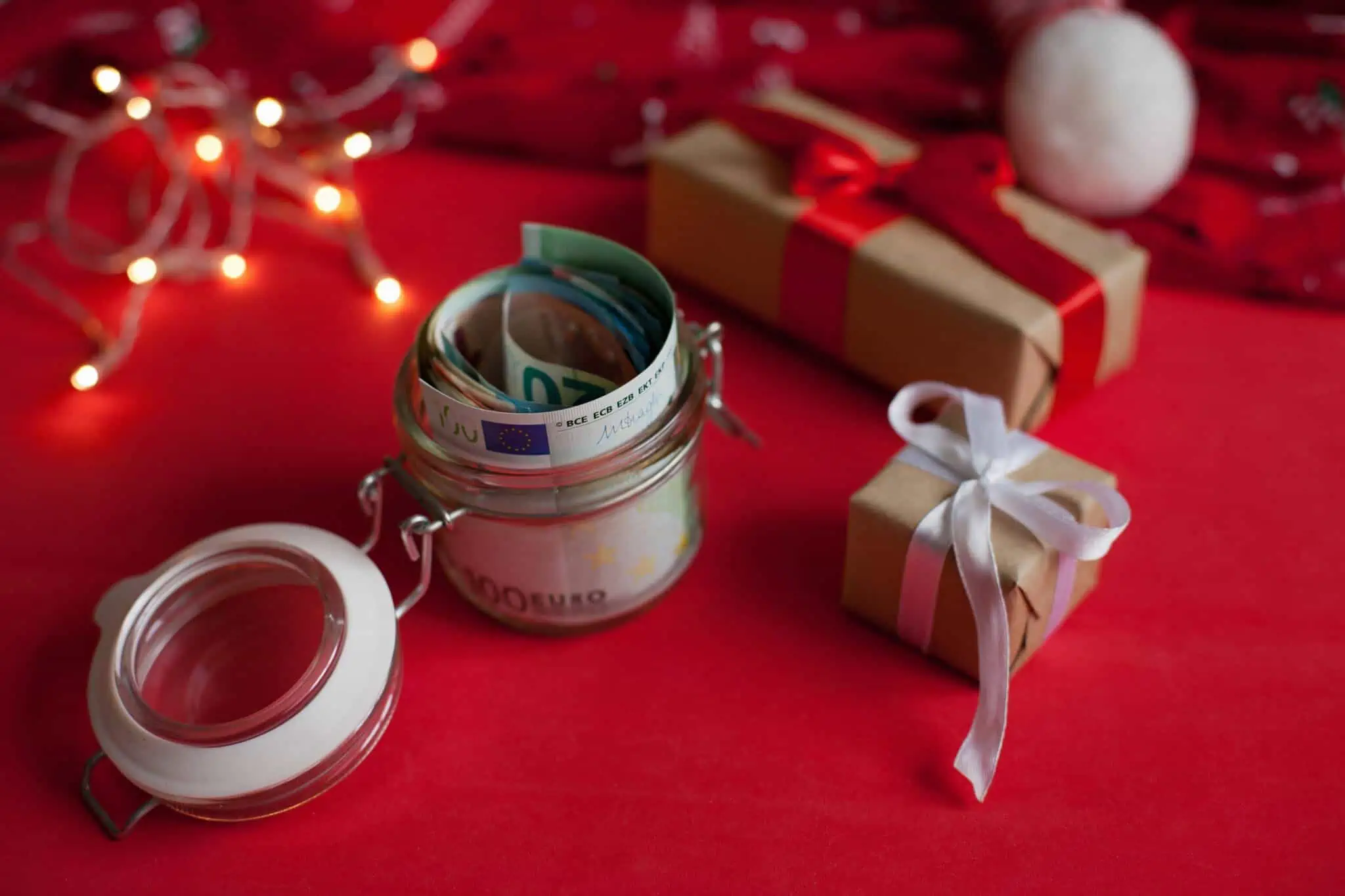Subsídio de Natal: 6 sugestões para a gestão do seu este ano numa mesa com toalha vermelha da quadra natalícia, por entre presentes e luzes, um frasco transparente com notas de euro ilustram o subsídio de Natal