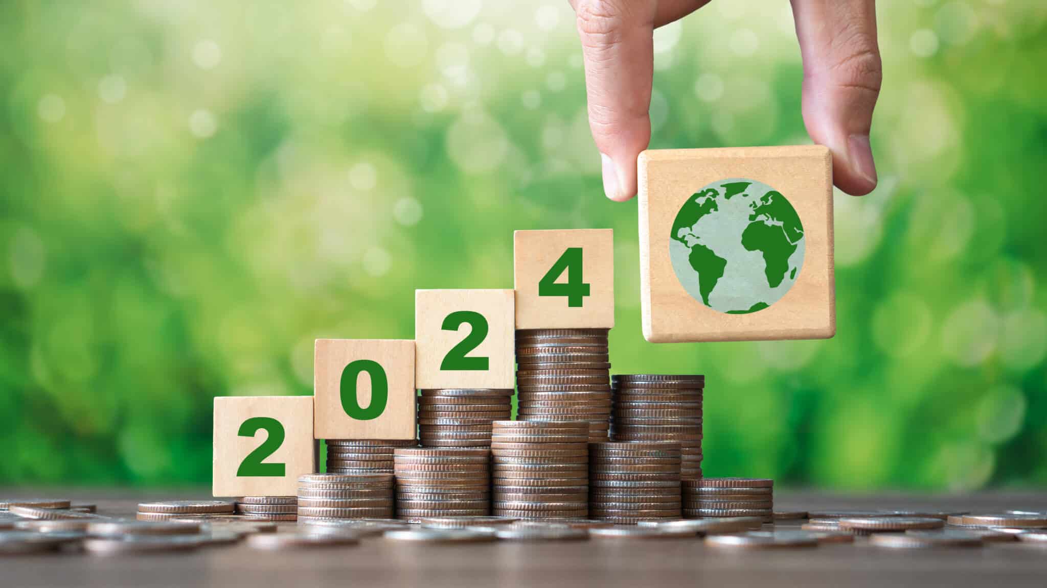 2024 mais sustentável: Dicas para poupar dinheiro e recursos em fundo verde de natureza desfocado, várias pequenas pilhas de moedas em crescendo têm uns cubos de madeira em cima mostrando o novo ano, finalizando com um cubo do planeta verde segurado por dois dedos, ilustram 2024 mais sustentável
