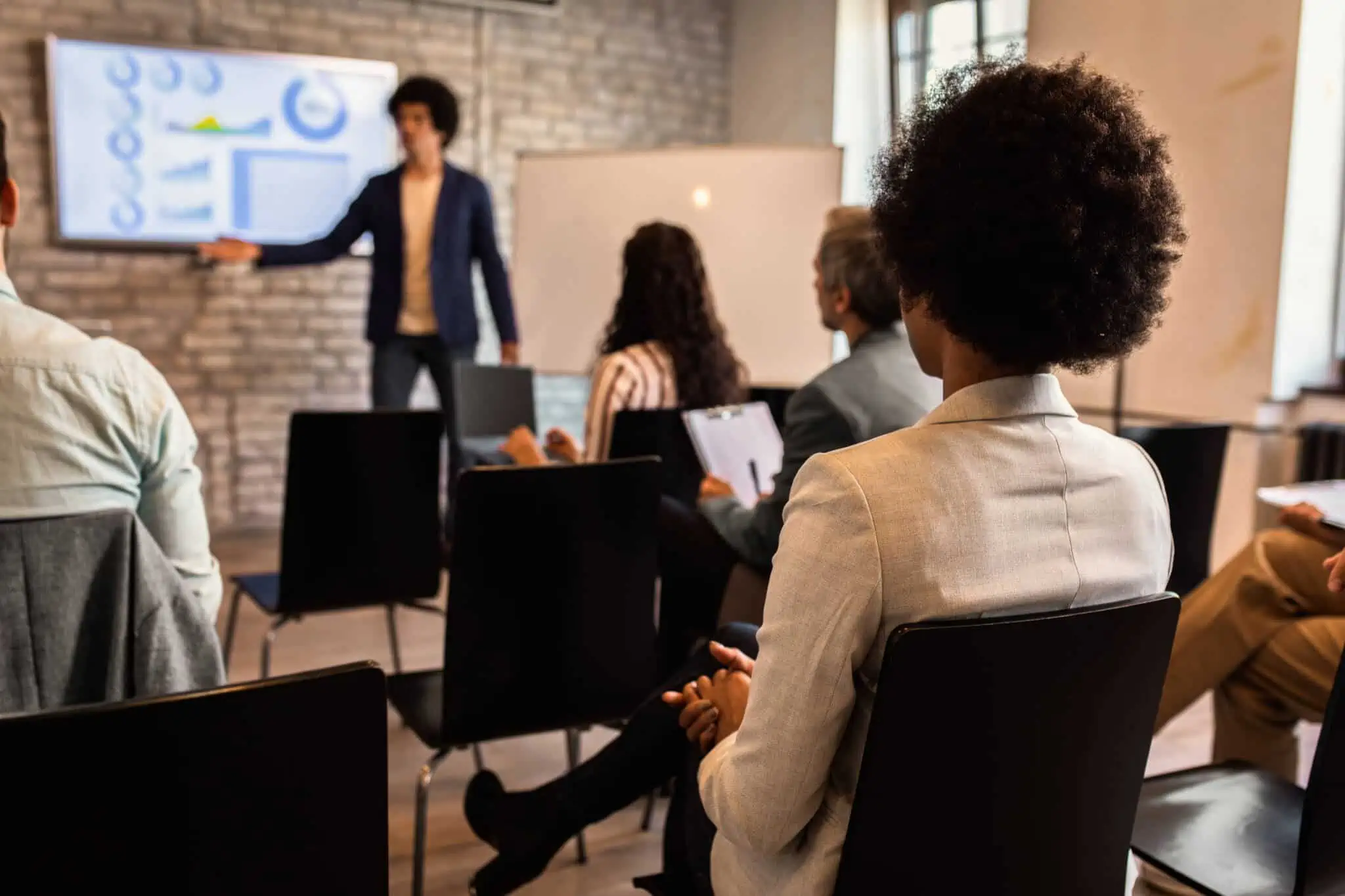 Certificação de Formadores: Como obter e quais os seus benefícios? jovem rapaz em fundo desfocado, dá formação a um conjunto de jovens sentados em cadeiras e mesas individuais, e para tal obteve certificação de formadores