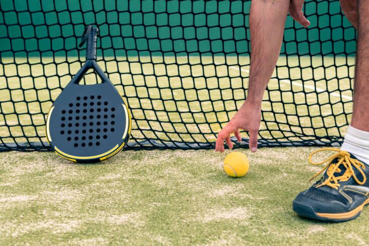 raquete e bola de padel encostados a uma rede de campo desta prática que deve obrigar a os praticantes terem seguros adequados
