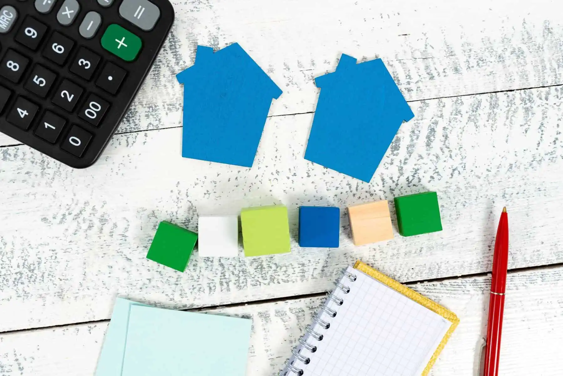 Sob folhas em branco, duas casinhas azuis, junto a uma calculadora e um bloco de notas e lápis, preparando as contas dos rendimentos prediais a constar do anexo IRS