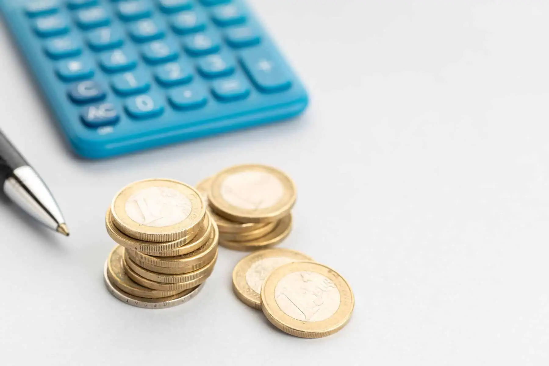 Quais os melhores investimentos com pouco dinheiro? Três molhos de moedas de euro sobre uma mesa, ao lado de uma calculadora azul e de uma caneta, numa alusão a investimentos com pouco dinheiro
