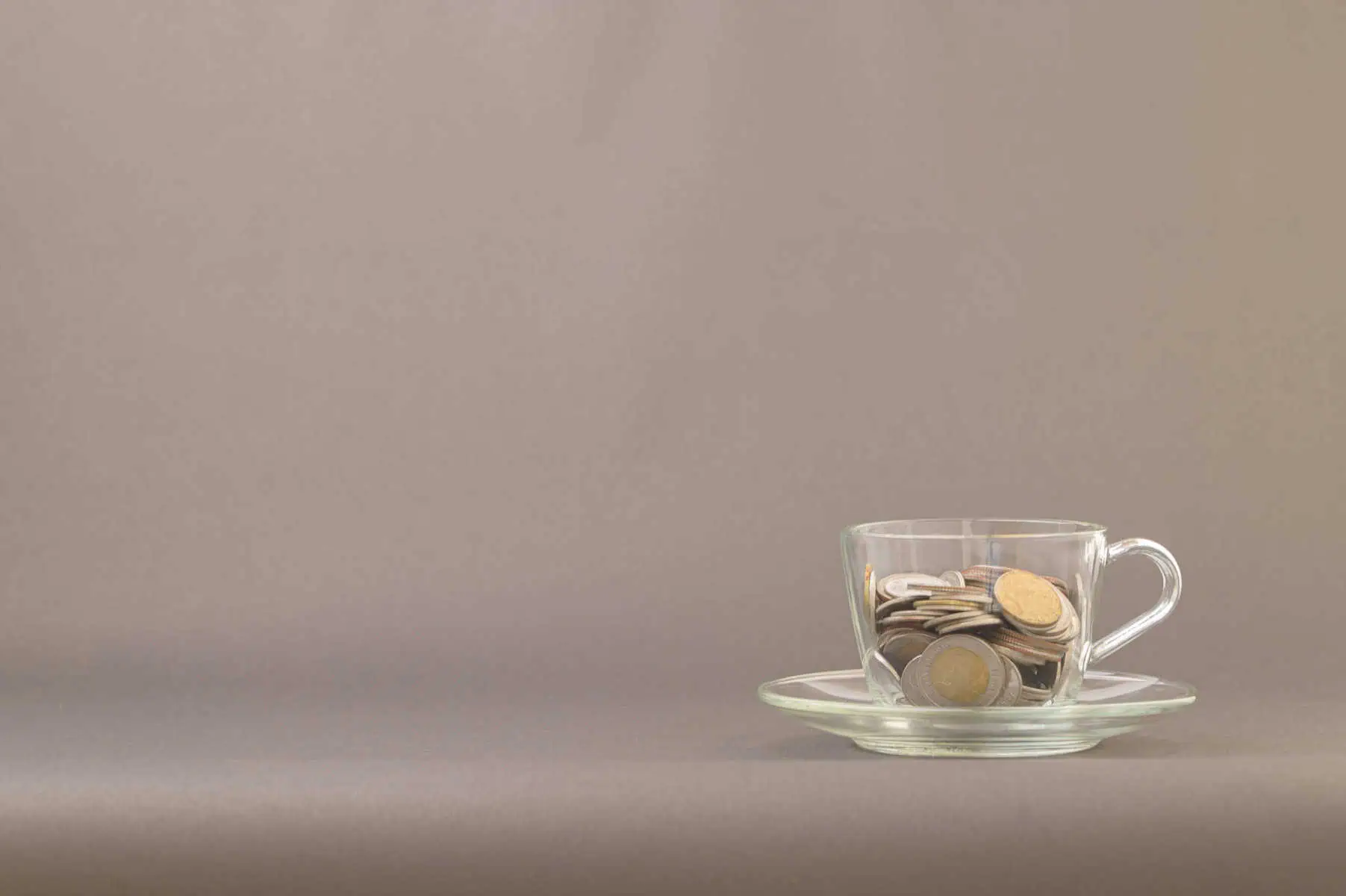 7 passos para começar a investir chavena de café de vidro transparente pousada em cima de um pires sobre uma mesa cinzenta. Dentro da chávena estão moedas de euro, numa alusão aos primeiros passos para começar a investir