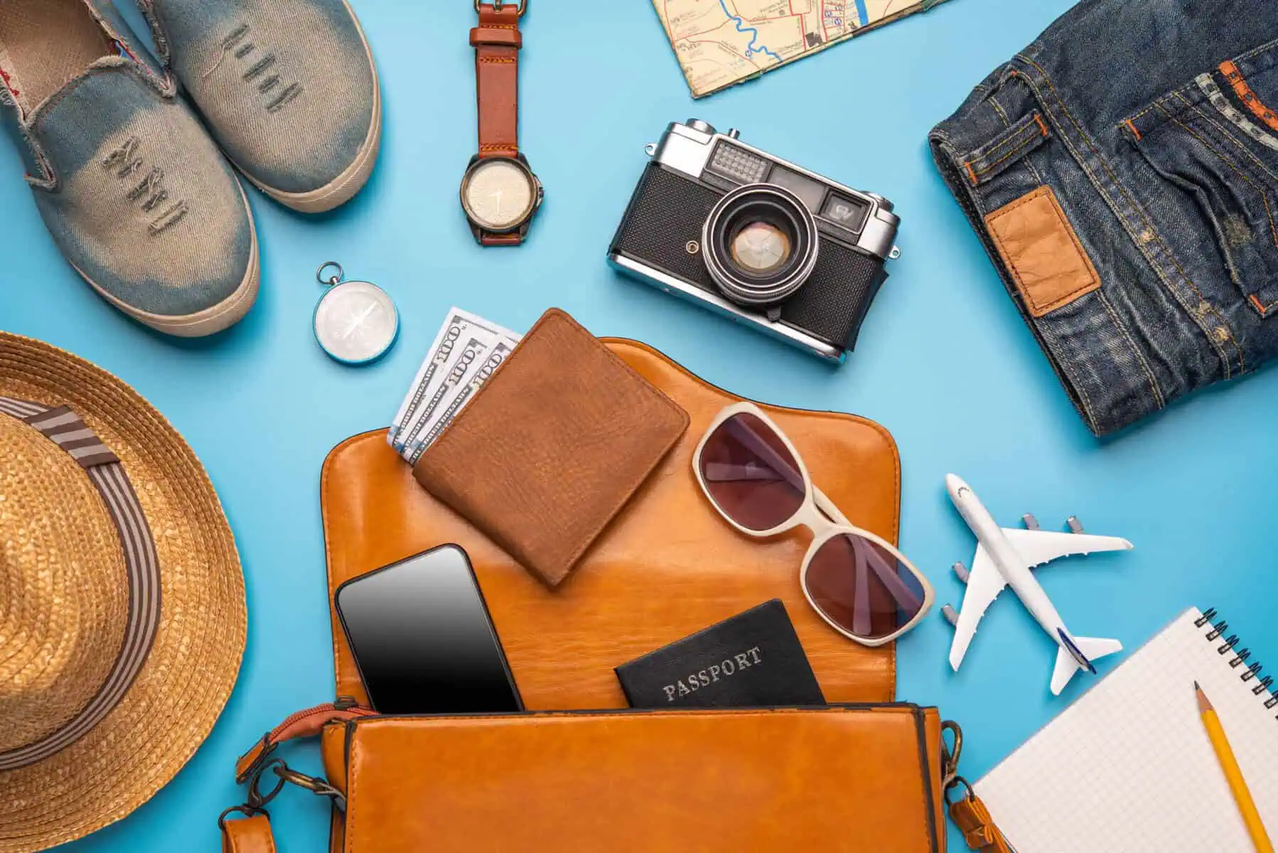 Seguro de viagem internacional: Qual a opção certa para o seu destino? carteira, óculos de sol, máquina fotográfica, roupa e sapatos, relógio e o passaporte espalhados em cima de uma mesa azul, numa alusão a uma viagem internacional