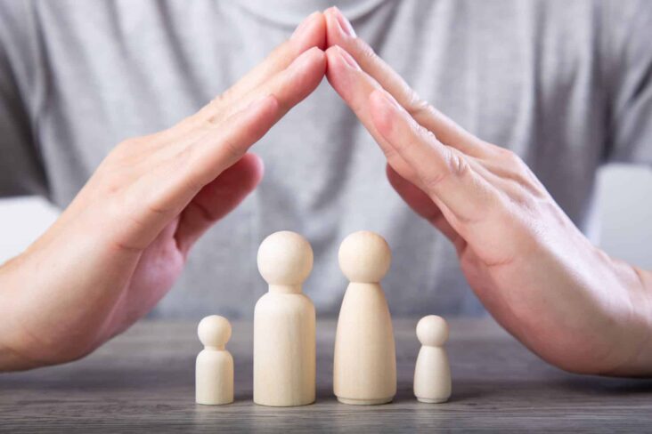 Mãos de homem, juntas a fazer a forma de telhado de uma casa. Em baixo estão quatro bonecos de madeira que simbolizam uma família. A fotografia remete-nos para seguros de vida