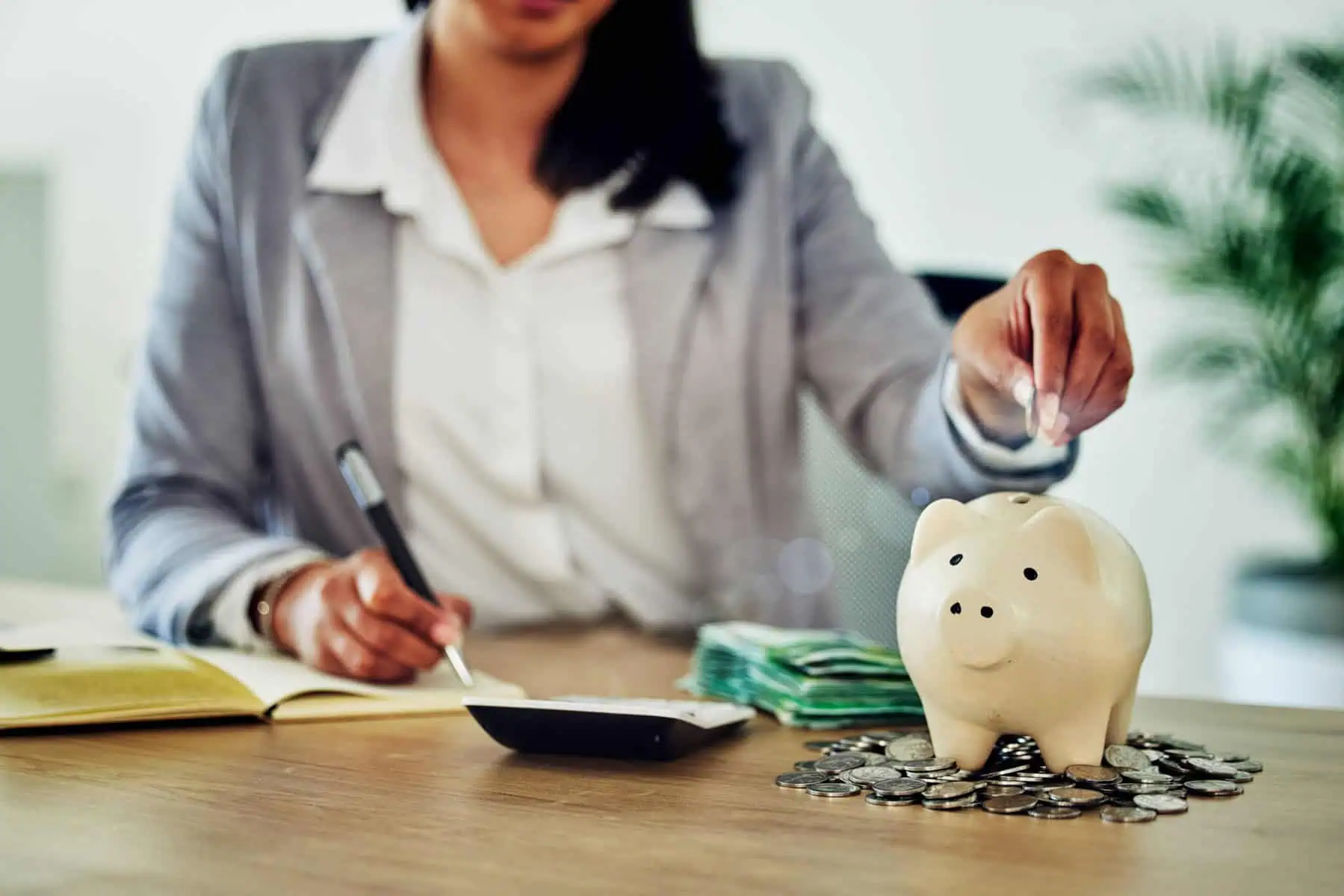 mulher sentada à secretária a tirar notas e a fazer contas com uma calculadora e um porquinho de mealheiro a ilustrar como organizar as suas finanças pessoais em 2025
