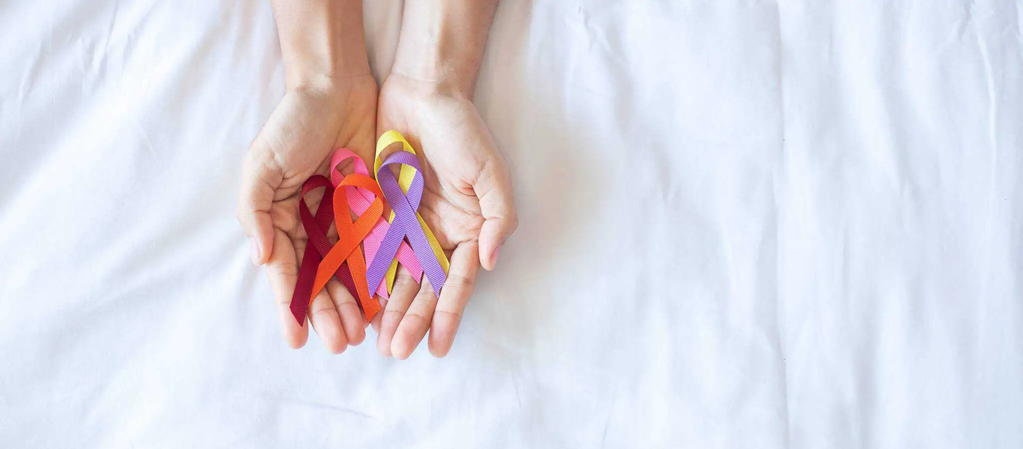 Qual a cobertura oncológica no seguro para doenças graves? Duas mãos de mulher juntas, em formato de concha, seguram laços de várias cores, numa alusão ao cancro