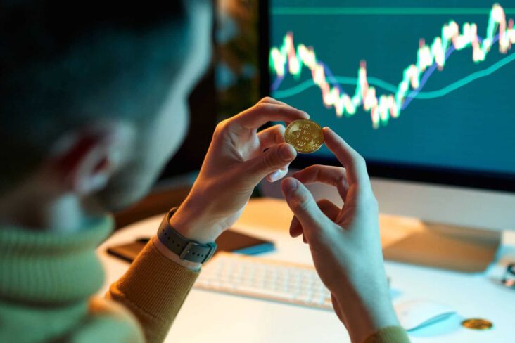 Investidor jovem olha para símbolo físico da bitcoin em frente a um computador com um gráfico de mercados financeiros.