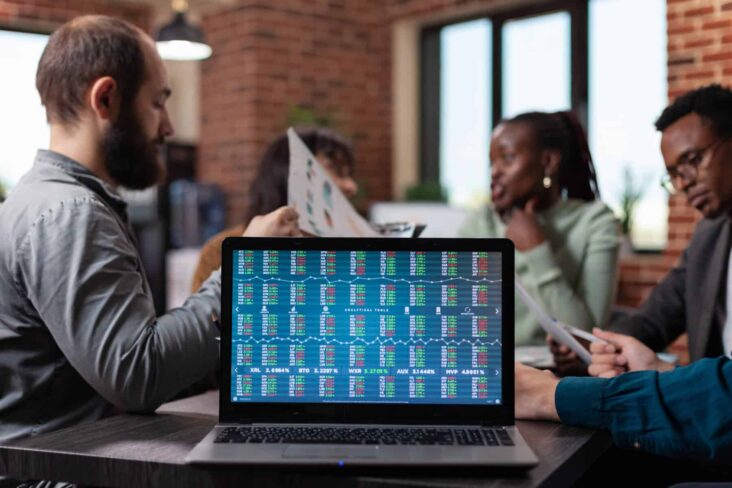 Computador ao centro de uma mesa, com tabelas onde se consegue ver o desempenho bolsista de vários ativos, entre os quais criptotativos. Por trás estão três pessoas a analisar o desempenho das suas carteiras.