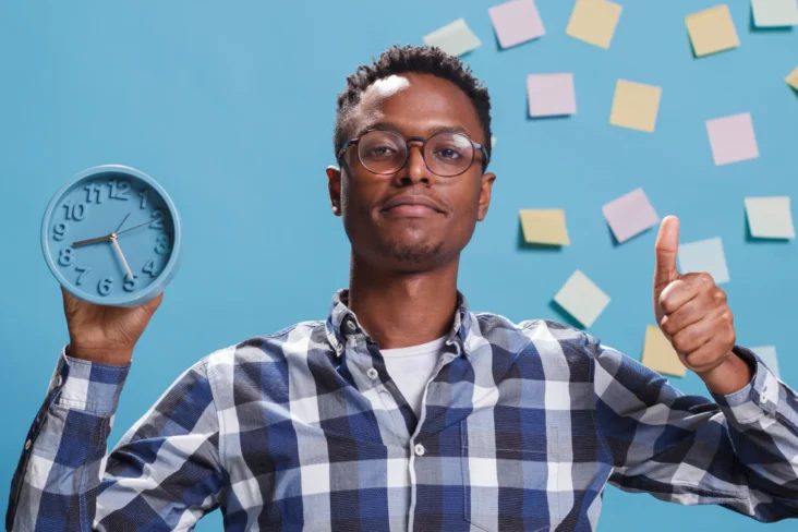 Homem segura em relógio à frente de uma parede azul repleta de notas de trabalho