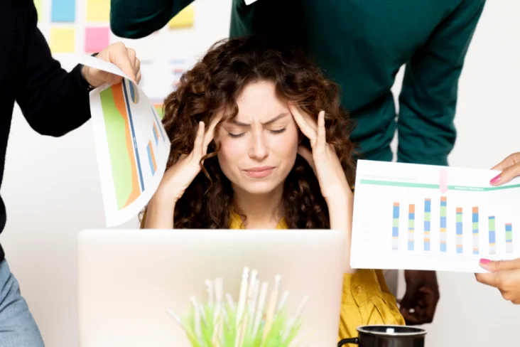 Mulher stressada no trabalho, sentada à secretária com vários colegas a pedirem-lhe mais trabalho