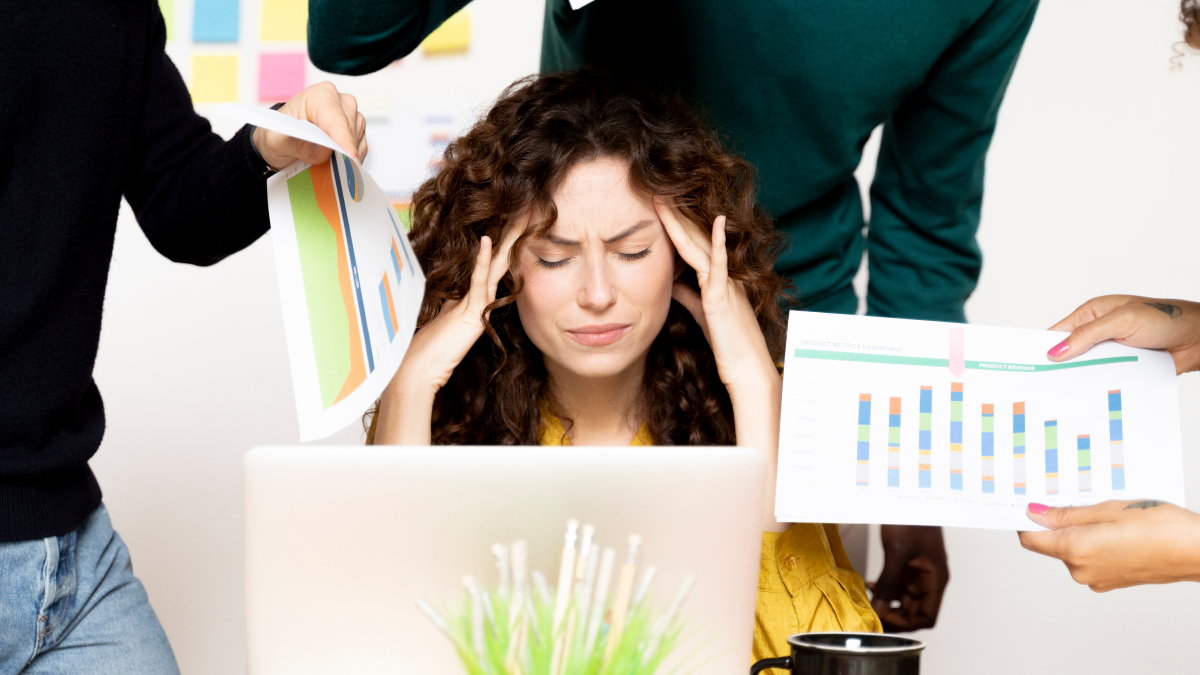 Mulher stressada no trabalho, sentada à secretária com vários colegas a pedirem-lhe mais trabalho