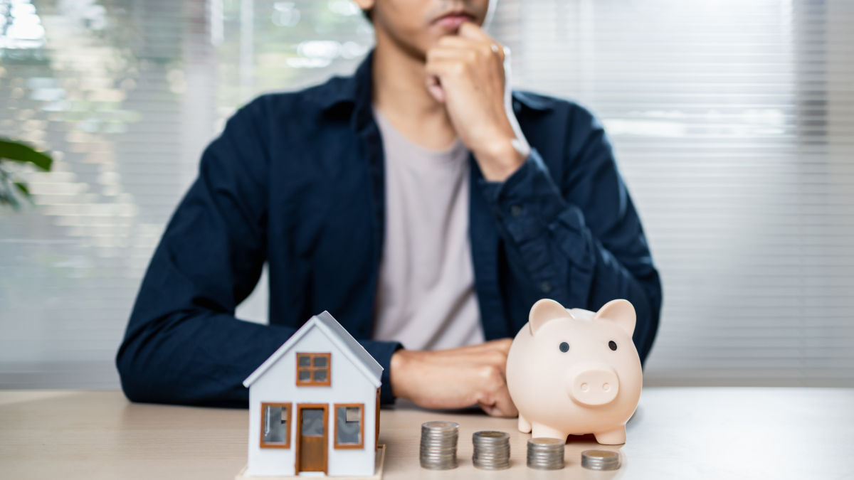 Homem a pensar sobre a transferência do crédito habitação, sentado à mesa, com uma casa de brincar, moedas e um porquinho mealheiro