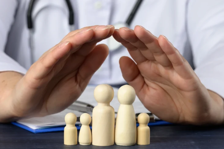 Imagem onde um médico protege com as mãos uma família de bonecos de madeira