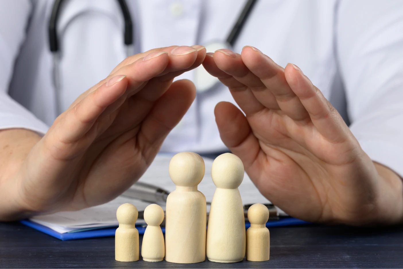 Os termos que deve conhecer antes de fazer um seguro de saúde Imagem onde um médico protege com as mãos uma família de bonecos de madeira