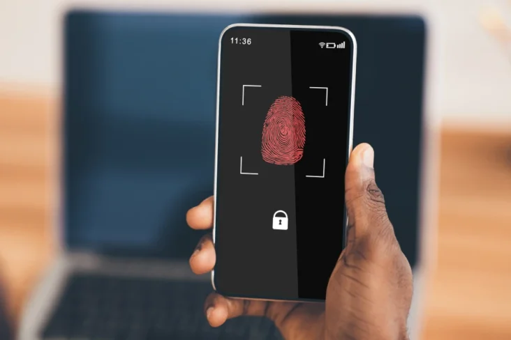 Imagem onde um homem segura num telemóvel em frente do seu computador, com um ecrão de autenticação de dois fatores (2FA)