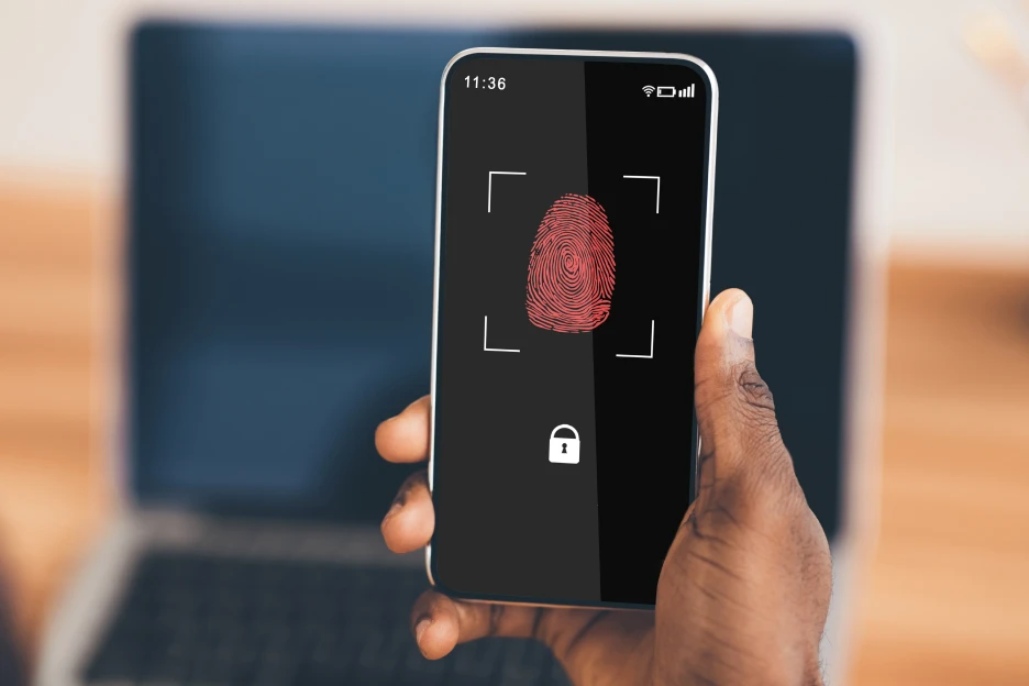 Autenticação multifator: Por que deve utilizá-la já na internet Imagem onde um homem segura num telemóvel em frente do seu computador, com um ecrão de autenticação de dois fatores (2FA)