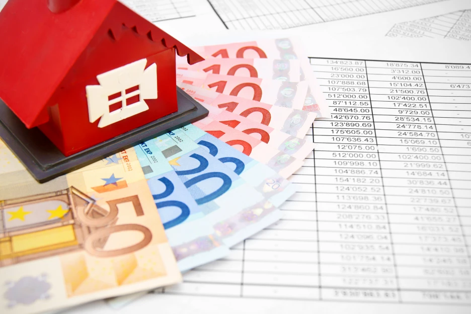 Escalões do IMT sobem 2% em 2026 e jovens ficam isentos até 330.539 euros Imagem de uma casa vermelha sobre dinheiro e uma tabela de escalões do IMT