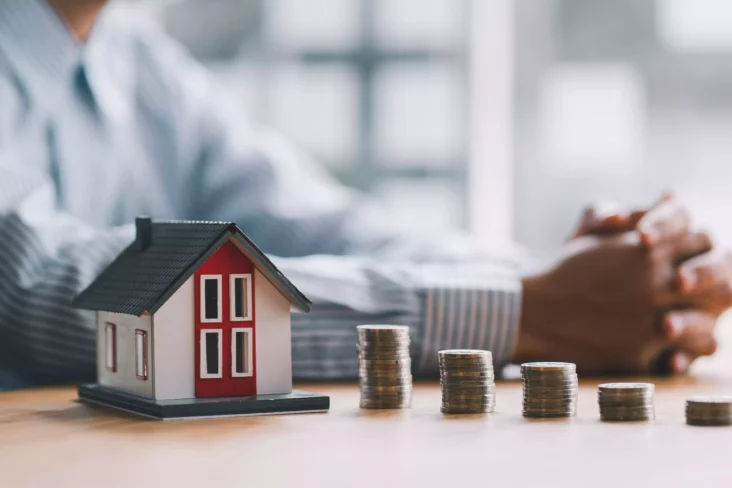 Homem junto a miniatura de casa e pilhas de moedas