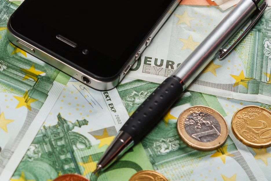 Como negociar o pacote de telecomunicações e evitar fidelizações abusivas Imagem de um telemovel sobre moedas e notas com uma caneta, representativo de contas de telecomunicações