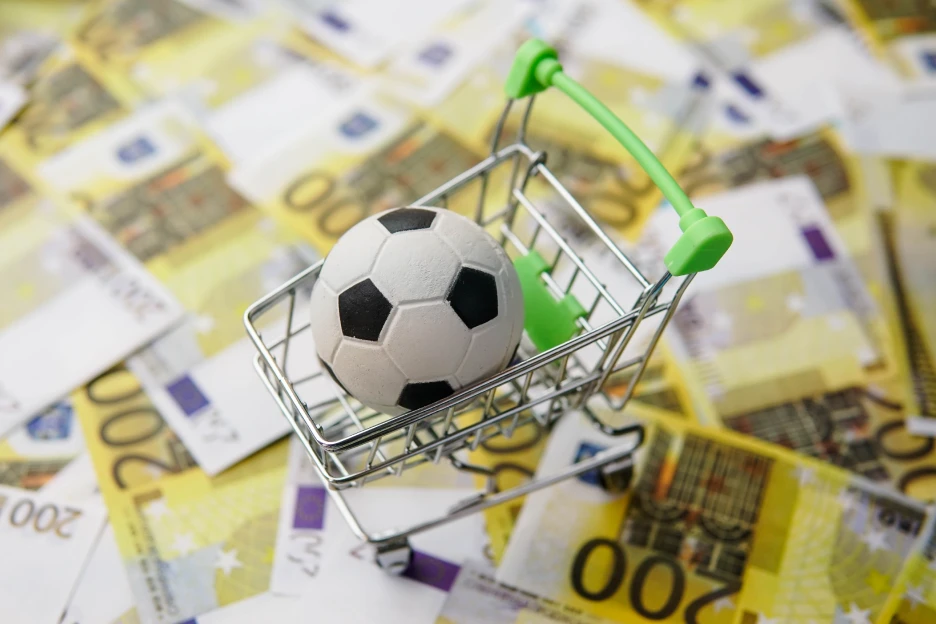 Quer comprar cromos do Mundial? Atenção às burlas Imagem de uma bola de futebol num carrinho de compras sobre notas, representativo do dinheiro que se pode gastar com futebol